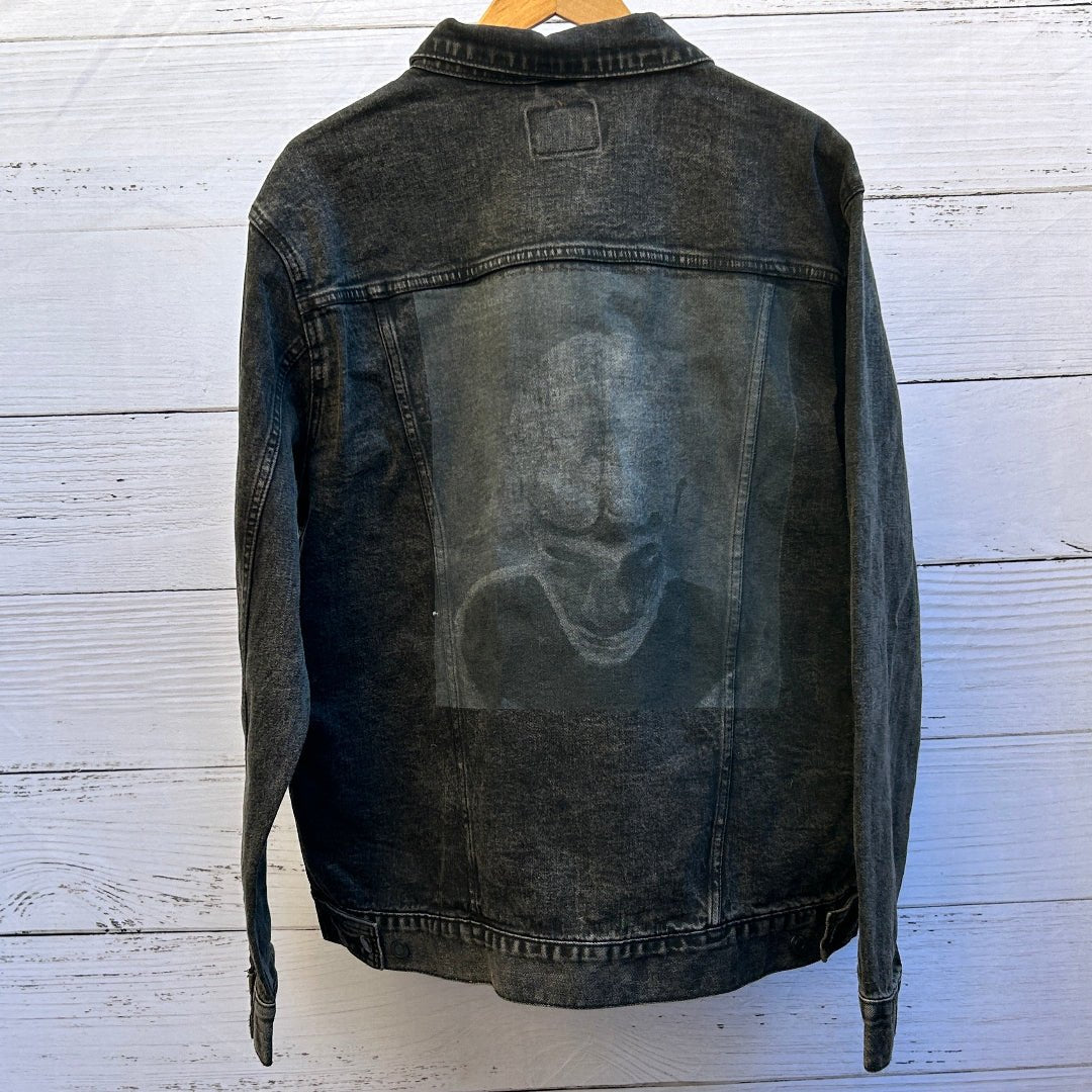 AS - IS. 21. Faded Monochrome Face. ONE - OF - ONE. Printed Denim Jacket. Large / Black Jacket custom denim jacket showing a faded monochrome face printed in soft gray tones, depicting a person with downturned eyes, heavy shadowing around the brow, nose, and mouth, and a ghostly, low-contrast appearance that blends into the denim; the artwork appears intentionally muted and worn, with the texture of the fabric and seams clearly visible around the rectangular print area, printed by DenimINK