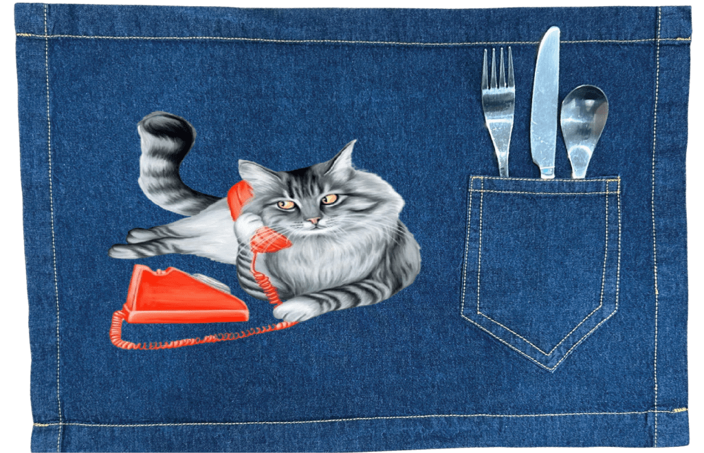 Mock up of a fluffy gray long-haired cat reclining with its tail curled upward, holding a bright red vintage telephone receiver to its ear Digitally printed by DenimINK on a Classic blue table placemat. the cat’s fur is rendered in layered charcoal &amp; silver tones with fine whisker lines and golden-yellow eyes gazing forward. a matching red phone base &amp; coiled cord rest below. titled “Cat on the Phone” &amp; created by Amélie Legault 