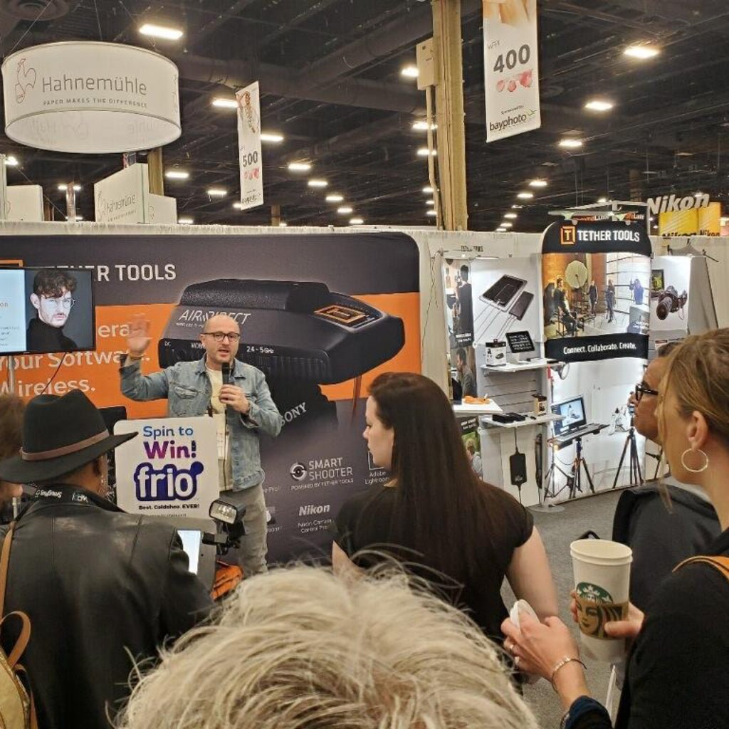 Back view of a speaker addressing a gathered crowd at a photography trade show while wearing a stonewashed denim jacket printed with “WASIO Photography.” Brand banners, lighting gear, and attentive faces surround the scene. The jacket becomes part of the presentation itself, carrying the photographer’s name and services into the noise, energy, and excitement of the show floor. Printed by DenimINK