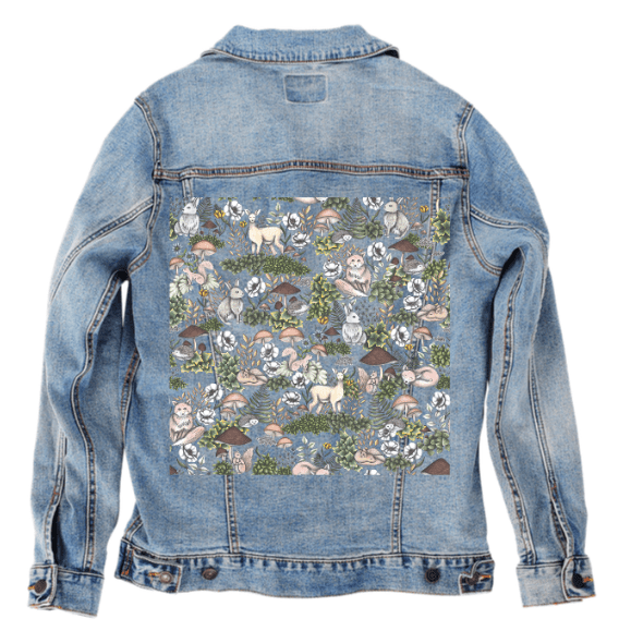 back view of a stonewashed denim jacket featuring the artwork “Into the Woods” by Cecilia Battaini, showing a richly layered woodland tapestry of deer, rabbits, owls, squirrels &amp; hedgehogs tucked between ferns, clovers, mushrooms &amp; ivy. the gentle fade of the denim enhances the earthy tones, giving the print a nostalgic, illustrated-fairytale feel. the design is digitally printed on the jacket’s back panel by DenimINK.