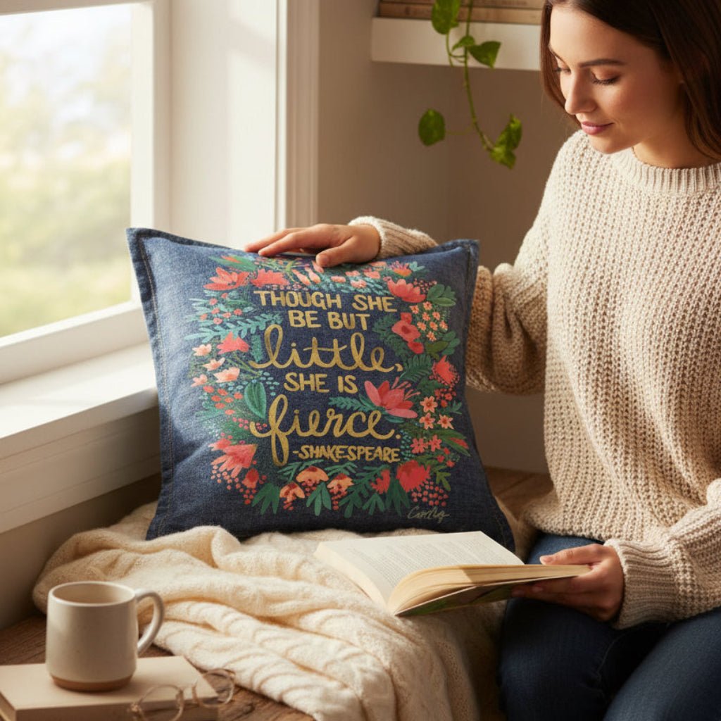 Woman reading a book with a decorative denim pillow featuring floral Shakespears text "Though She Be But Little, She is Fierce" . little-and-fierce-by-CatCoq-denim-throw-pillow-lifestyle-shot