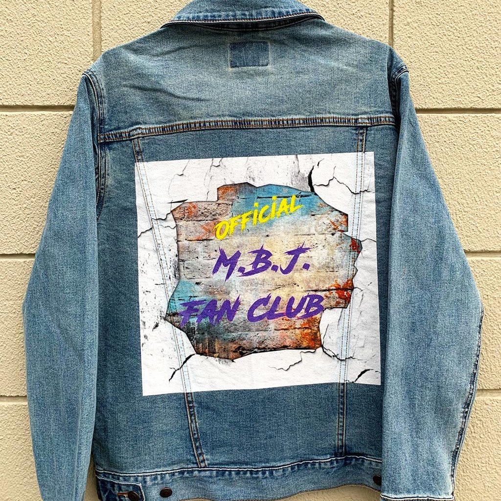 Back view of a stonewashed denim jacket hanging against a pale brick wall, featuring a rectangular artwork that looks torn through plaster to reveal layered texture beneath. The words “OFFICIAL M.B.J. FAN CLUB” appear in bold yellow and purple, surrounded by cracked edges and weathered tones of rust, teal, and concrete white, as if the jacket carries the echo of underground shows and fan devotion pressed into denim.