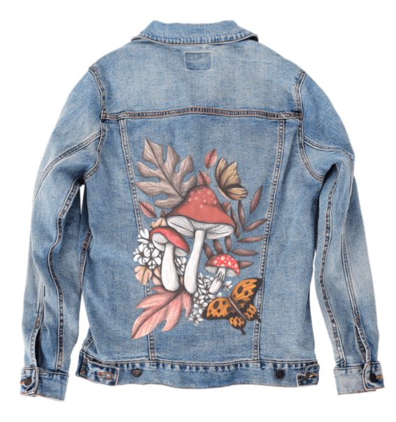 back view of a stonewashed denim jacket featuring a soft botanical composition of red mushrooms with white speckles, wildflowers, faded oak leaves &amp; a golden butterfly, drawn in pencil textures &amp; watercolor tones. the weathered denim background gives the artwork an earthy, enchanted forest feel. the design, “Mushroom Magic 2” by Cecilia Battaini,is digitally printed on the jacket’s back panel by DenimINK.