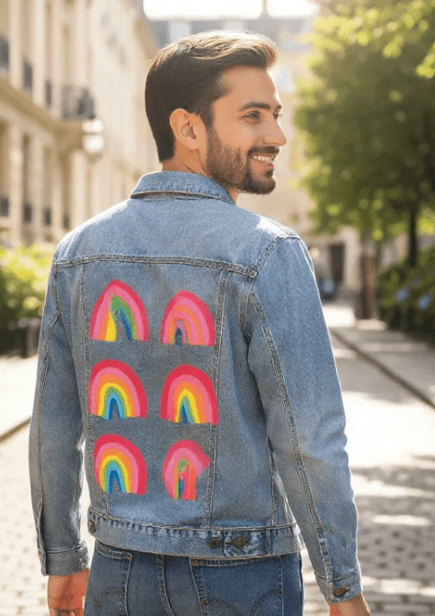 three-quarter rear view of a smiling model on a cobblestone street wearing a stonewashed denim jacket printed with the “Rainbow Collection” by CatCoq. the design shows six hand-painted rainbows arranged in two columns, each with vibrant arcs of red, orange, yellow, green, blue &amp; purple. sunlight highlights the texture of the watercolor artwork. printed by DenimINK.