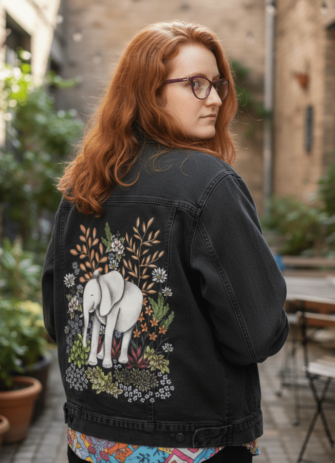 back view of a model wearing a black denim jacket in a quiet alley café, surrounded by potted plants. the jacket’s back panel displays a whimsical white elephant nestled in a blooming botanical jungle of peachy leaves, white daisies, lavender blooms, &amp; wild greenery. the artwork “Safari Elephant” by Cecilia Battaini is digitally printed on the jacket’s back panel by DenimINK.