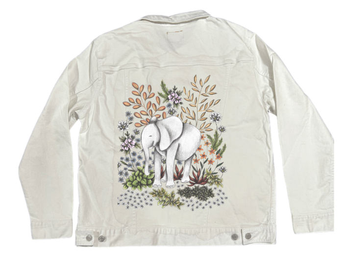 back view of a white denim jacket featuring a gentle safari-themed design of a white elephant nestled among stylized botanicals in pastel lavender, pale gold, ivory &amp; mossy green. the soft contrast between the flora &amp; fauna gives the artwork a quiet, organic feel. the artwork “Safari Elephant” by Cecilia Battaini is digitally printed on the jacket’s back panel by DenimINK.