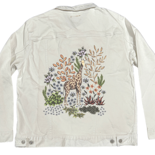 back view of a white denim jacket mockup featuring a printed safari-style illustration of a giraffe standing in a mix of dusty orange foliage, trailing white blooms, &amp; olive-green desert plants. the bold black canvas contrasts the delicate pencil strokes &amp; muted colors. the artwork “Safari Giraffe” by Cecilia Battaini is digitally printed on the jacket’s back panel by DenimINK.