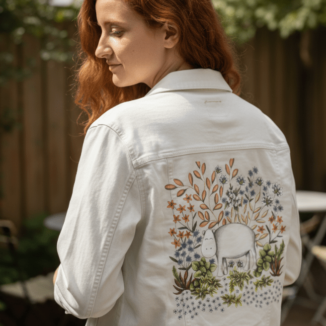 three-quarter rear view of a model standing near a sun-dappled garden patio, wearing a white denim jacket featuring a gently smiling white hippo among warm brown branches, soft lavender blossoms, blue &amp; white wildflowers, &amp; succulent greenery. the artwork “Safari Hippo” by Cecilia Battaini is digitally printed on the jacket’s back panel by DenimINK.