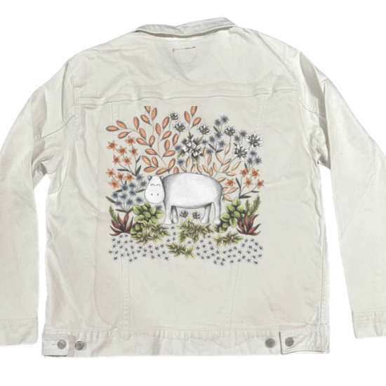 back view of a white denim jacket featuring a cheerful white hippo in the center of an ornate garden illustration filled with pastel botanicals, warm clay-toned leaves, soft purples, ivory flowers &amp; desert greens. the artwork “Safari Hippo” by Cecilia Battaini is digitally printed on the jacket’s back panel by DenimINK.
