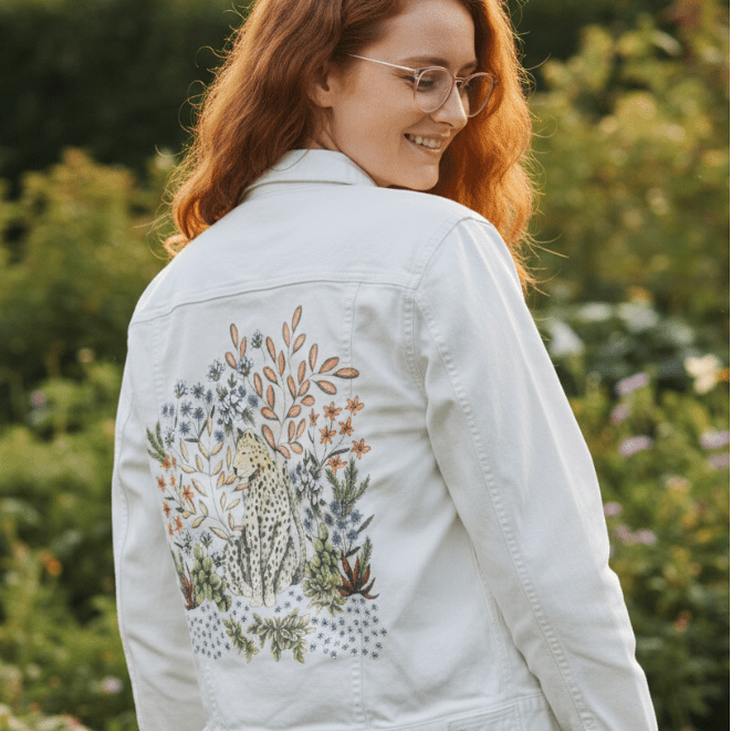 three-quarter rear view of a model standing in a lush garden full of greenery &amp; late-summer flowers, smiling while wearing a white denim jacket with a soft-toned botanical artwork on the back. the design features a relaxed leopard nestled among pale lavender flowers, clay-orange leaves, &amp; mint succulents. sunlight warms the scene, making the printed illustration glow softly against the crisp white denim. the artwork “Safari Leopard” by Cecilia Battaini is digitally printed on the jacket’s back panel by Deni