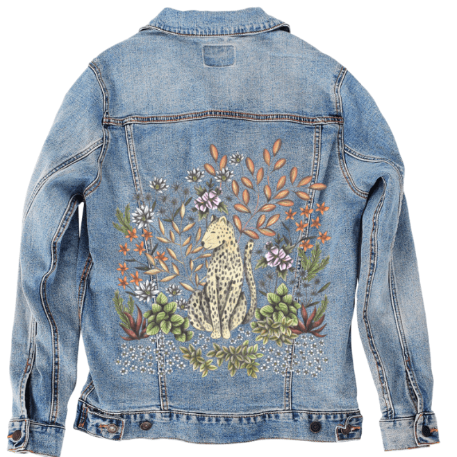 back view of a stonewashed denim jacket mockup featuring a gentle jungle composition showing a spotted leopard seated in a dense bed of tropical foliage &amp; soft wildflowers. pale green succulents, blue daisies, coral blossoms, &amp; creamy leaves wrap around the feline figure with a vintage, worn-in look against the faded blue fabric. the artwork “Safari Leopard” by Cecilia Battaini is digitally printed on the jacket’s back panel by DenimINK.