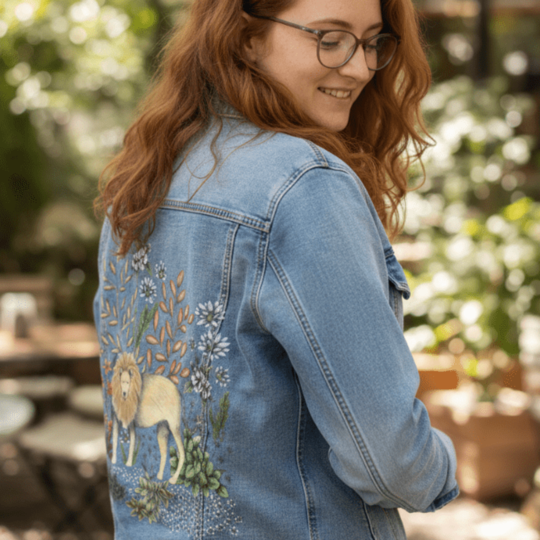 three-quarter rear view of a model wearing a stonewashed denim jacket with a digitally printed lion surrounded by softly drawn flowers, leafy stems, and textured greenery. the setting is a peaceful café garden, with filtered light highlighting the jacket’s muted tones. the artwork “Safari Lion” by Cecilia Battaini is digitally printed on the jacket’s back panel by DenimINK.