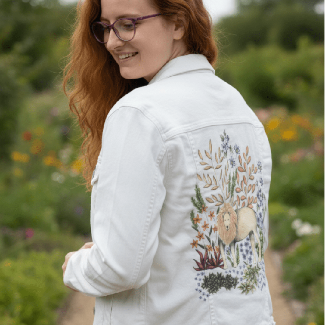 three-quarter rear view of a model walking through a wildflower-filled path wearing a white denim jacket. the digitally printed back panel features a golden-maned lion standing calmly amidst pale daisies, orange blossoms, desert leaves, and sage-toned succulents. the muted gray sky gives the floral scene a soft, dreamlike quality. the artwork “Safari Lion” by Cecilia Battaini is digitally printed on the jacket’s back panel by DenimINK.