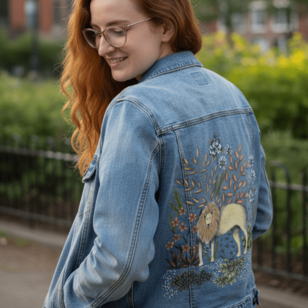 three-quarter rear view of a model in a stonewashed denim jacket smiling near a black iron fence lined with soft green bushes. the back panel features a printed illustration of a golden lion surrounded by warm rust-colored leaves, succulents, and clusters of white flowers. the artwork “Safari Lion” by Cecilia Battaini is digitally printed on the jacket’s back panel by DenimINK.