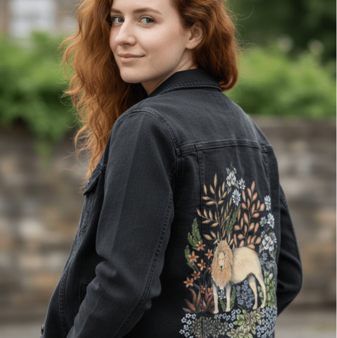 three-quarter rear view of a model standing near a pale stone wall while wearing a black denim jacket. the printed back panel features a regal lion in soft golden tones, surrounded by botanical elements—amber leaves, white wildflowers, mossy succulents, and fine pencil shading throughout. the artwork “Safari Lion” by Cecilia Battaini is digitally printed on the jacket’s back panel by DenimINK.