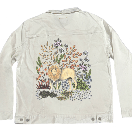 back view of a white denim jacket mockup featuring a digitally printed illustration of a golden-maned lion nestled in layered botanical surroundings. the crisp white denim highlights the subtle pencil textures &amp; soft pastel tones of the surrounding succulents, blossoms, &amp; fern-like leaves. the artwork “Safari Lion” by Cecilia Battaini is digitally printed on the jacket’s back panel by DenimINK.