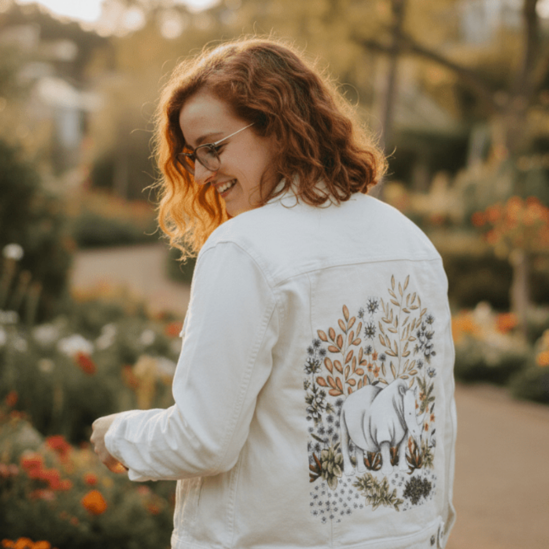 three-quarter rear view of a model walking through a late afternoon flower garden in a white denim jacket featuring a softly drawn white rhino standing among wild blooms and succulents. the backdrop includes golden branches, pale purple and orange blossoms, and detailed pencil-shaded foliage. the artwork “Safari Rhino” by Cecilia Battaini is digitally printed on the jacket’s back panel by DenimINK.