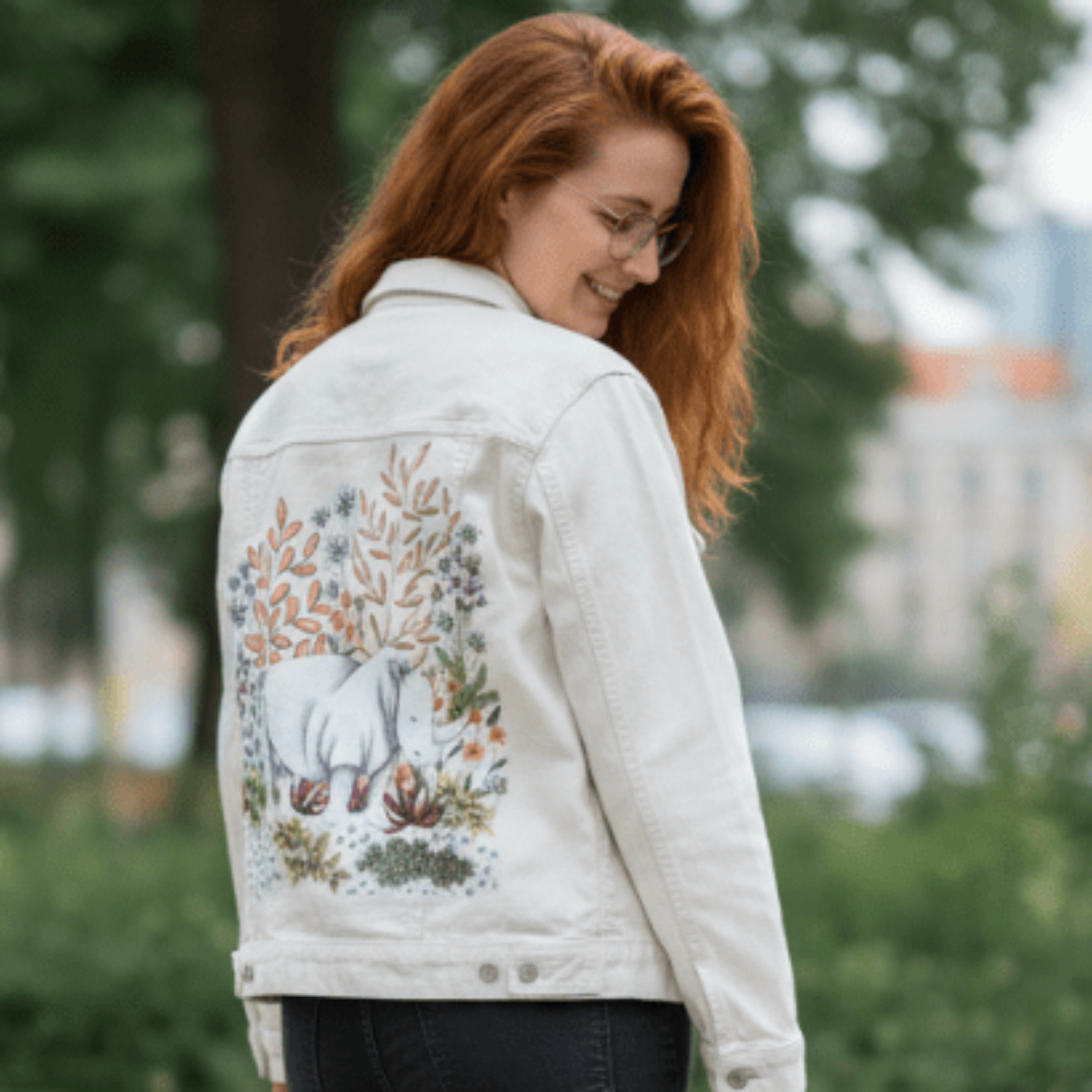 rear view of a smiling model wearing a white denim jacket with an illustration of a white rhino nestled in a lush forest of hand-drawn foliage and wildflowers. warm coral and terracotta tones blend with deep green leaves and pale florals. the artwork “Safari Rhino” by Cecilia Battaini is digitally printed on the jacket’s back panel by DenimINK.