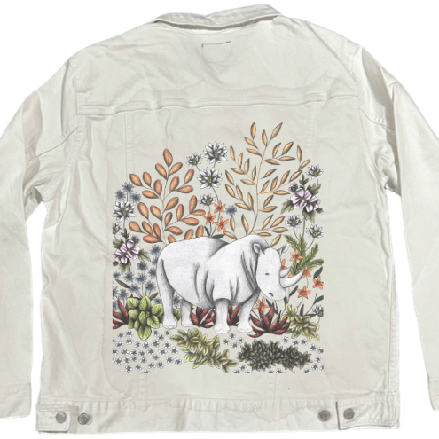 back view of a white denim jacket mockup printed with a delicate forest-meets-desert illustration of a white rhino standing among sketched blossoms, soft green succulents, and sienna-toned leaves. the artwork “Safari Rhino” by Cecilia Battaini is digitally printed on the jacket’s back panel by DenimINK.