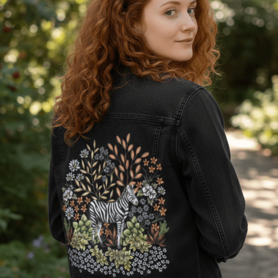 back view of model standing on a shaded woodland path wearing a black denim jacket featuring the “Safari Zebra” artwork by Cecilia Battaini. the illustration is digitally printed on the back panel and features a zebra surrounded by dense foliage—creamy blossoms, golden leaves, soft blue wildflowers, and lush succulents—arranged in a half-circle that glows against the dark denim. printed by DenimINK.
