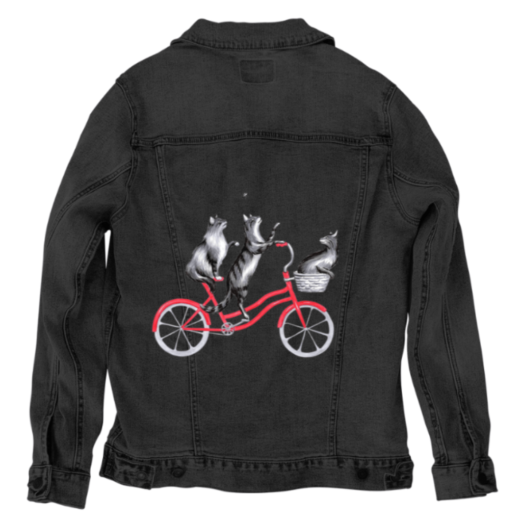 back view of a black denim jacket featuring the artwork “Cats on a Bicycle” by Amélie Legault, digitally printed on the back panel by DenimINK. the illustration shows two gray cats riding together on a red bicycle, their fur softly textured and whimsical against the worn denim grain. sunlight grazes the jacket seams, highlighting contrast stitching and metal rivets. the artwork is digitally printed on the jacket’s back panel by DenimINK