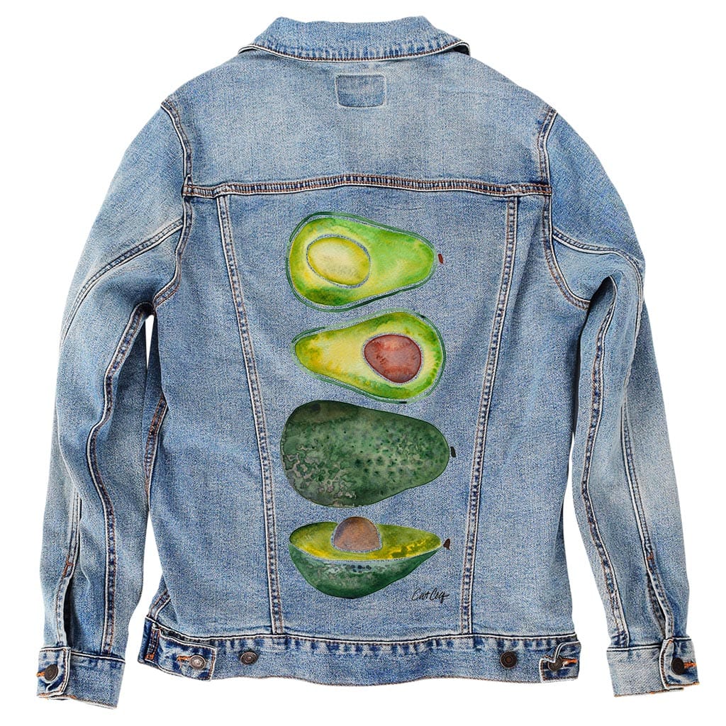 back view of a stonewashed denim jacket featuring the artwork “Avocado Slices” by CatCoq, digitally printed on the back panel. the vertically stacked composition includes two avocado halves—one with pit, one without—a full avocado with pebbled skin, and another halved slice. the washed denim softens the contrast, giving the green tones a slightly muted appearance while retaining the texture of the watercolor.