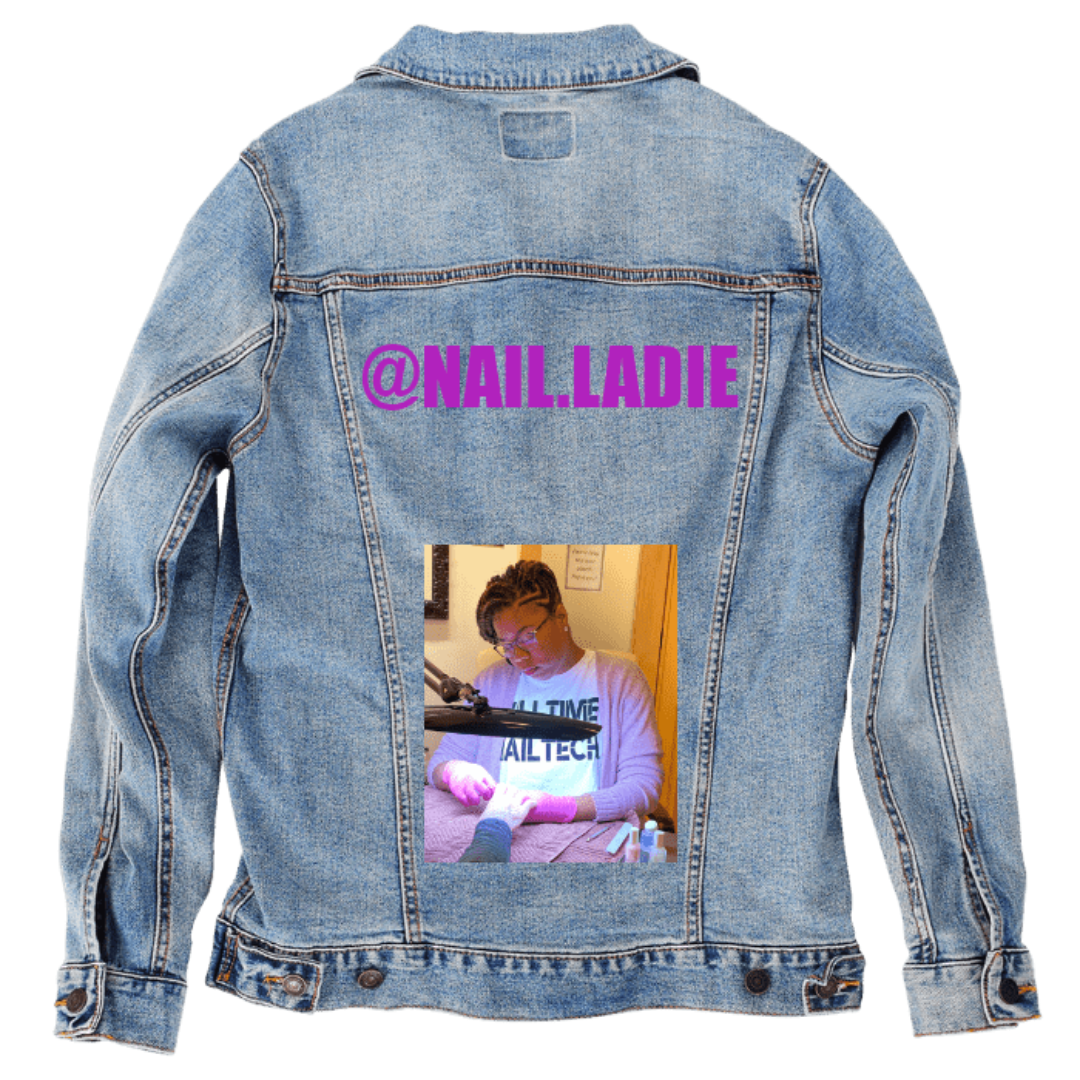 Customer designed custom denim jacket (M stonewash) featuring bold purple lettering across the upper back that reads “@NAIL.LADIE,” paired with a photo below of a nail technician wearing gloves while working under a bright task lamp. The warm indoor lighting, detailed hand-painting setup, and personalized handle text create a vibrant, professional, and expressive design that stands out against the faded blue denim, printed on denim jacket by DenimINK.