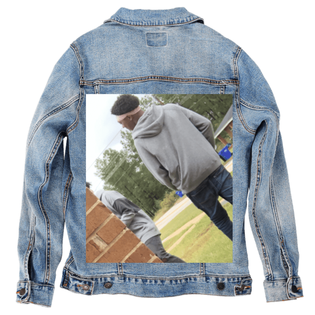 Customer designed custom denim jacket (M stonewash) featuring an outdoor candid photo printed on the back, showing a person wearing a gray hoodie and jeans walking near a brick wall, with another partially visible figure in the foreground. The background includes grassy areas, trees, and a roadway, giving the image a natural, everyday setting. The personal snapshot contrasts with the soft, faded blue denim, creating a relaxed custom design, printed on denim jacket by DenimINK.