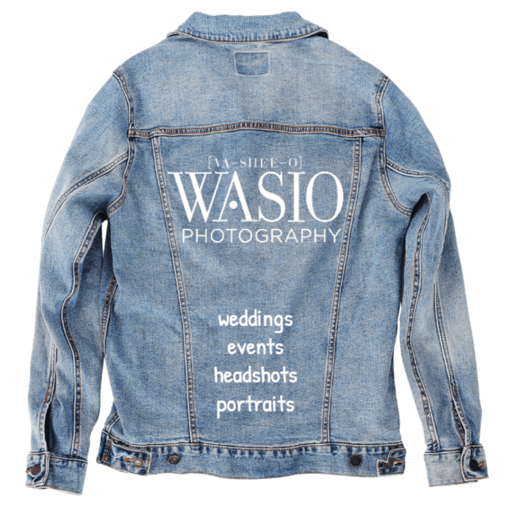 Back view of a stonewashed denim jacket shown as a studio mockup, featuring clean white typography reading “In the style of WASIO Photography” across the upper back. A vertical list below spells out services including weddings, events, headshots, and portraits. The design feels crisp and professional, like a walking business card, with the texture of denim grounding the polished branding in something wearable and real. Printed by DenimINK