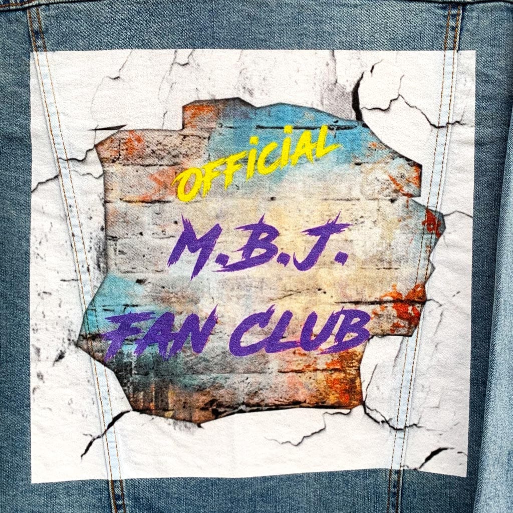 Close-up view of the “OFFICIAL M.B.J. FAN CLUB” artwork printed on the back of a stonewashed denim jacket. Cracked white edges frame a torn-wall effect revealing layered rust, teal, and concrete textures beneath bold yellow and purple lettering. The surface feels gritty and tactile, like paint pressed into fabric, carrying the weight of fan loyalty, late-night music, and walls covered in history.