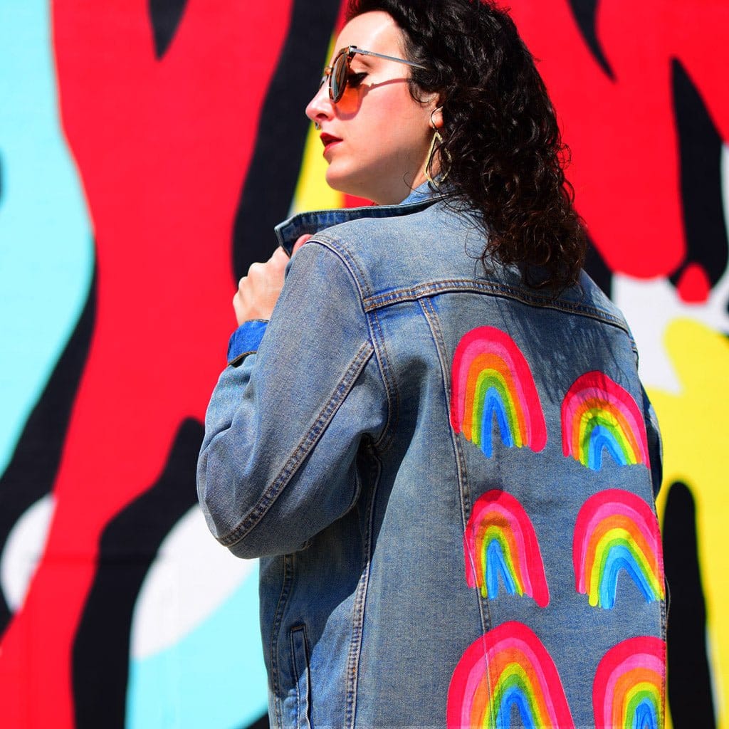 side view of a model in front of a bold, graphic mural wall wearing a denim jacket printed with the “Rainbow Collection” by CatCoq. the six rainbow arches stand out against the jacket’s stonewashed texture, contrasting with the vivid red, blue &amp; yellow background behind them. the model lifts the collar slightly with one hand. printed by DenimINK.