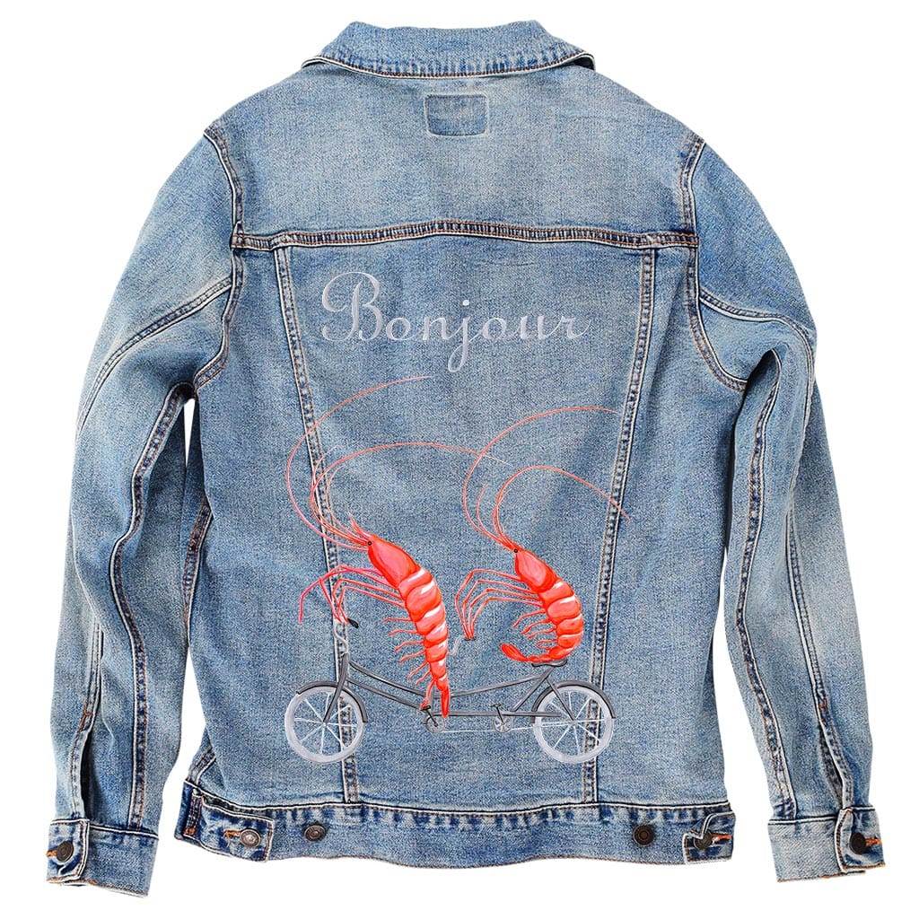 back view of a stonewash denim jacket featuring the artwork “Shrimp Riding a Bicycle” by Amélie Legault, showing two red shrimp pedaling a small bicycle beneath the word “Bonjour.” the artwork is digitally printed on the jacket’s back panel by DenimINK