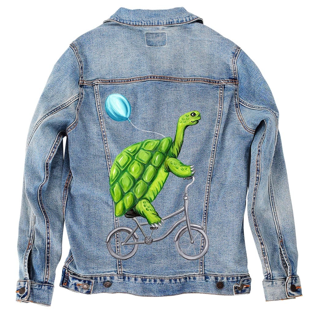 back view of a stonewash denim jacket featuring the artwork “Turtle Riding a Bicycle” by Amelia Legault, showing a green turtle riding a bicycle with a blue balloon tied to its shell. the artwork is digitally printed on the jacket’s back panel by DenimINK